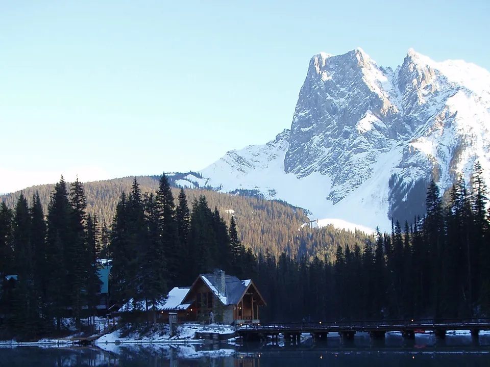 A wooden lodge and bridge sit by a dark lake with snow-capped mountains and pine trees in the background.
