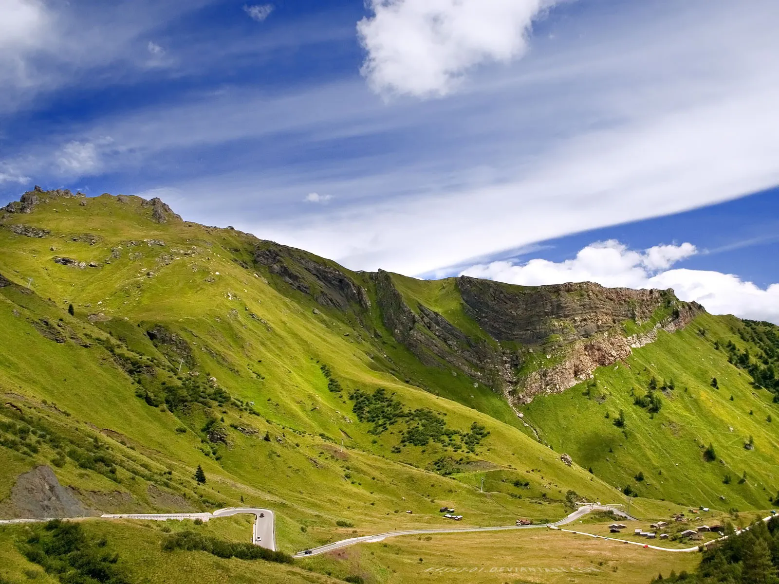 A winding road curves through vibrant green mountain slopes under a blue sky with wispy clouds.