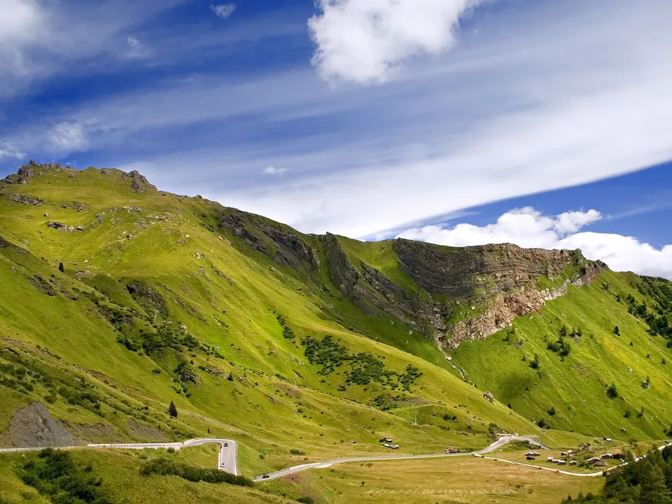 A winding road curves through vibrant green mountain slopes under a blue sky with wispy clouds.