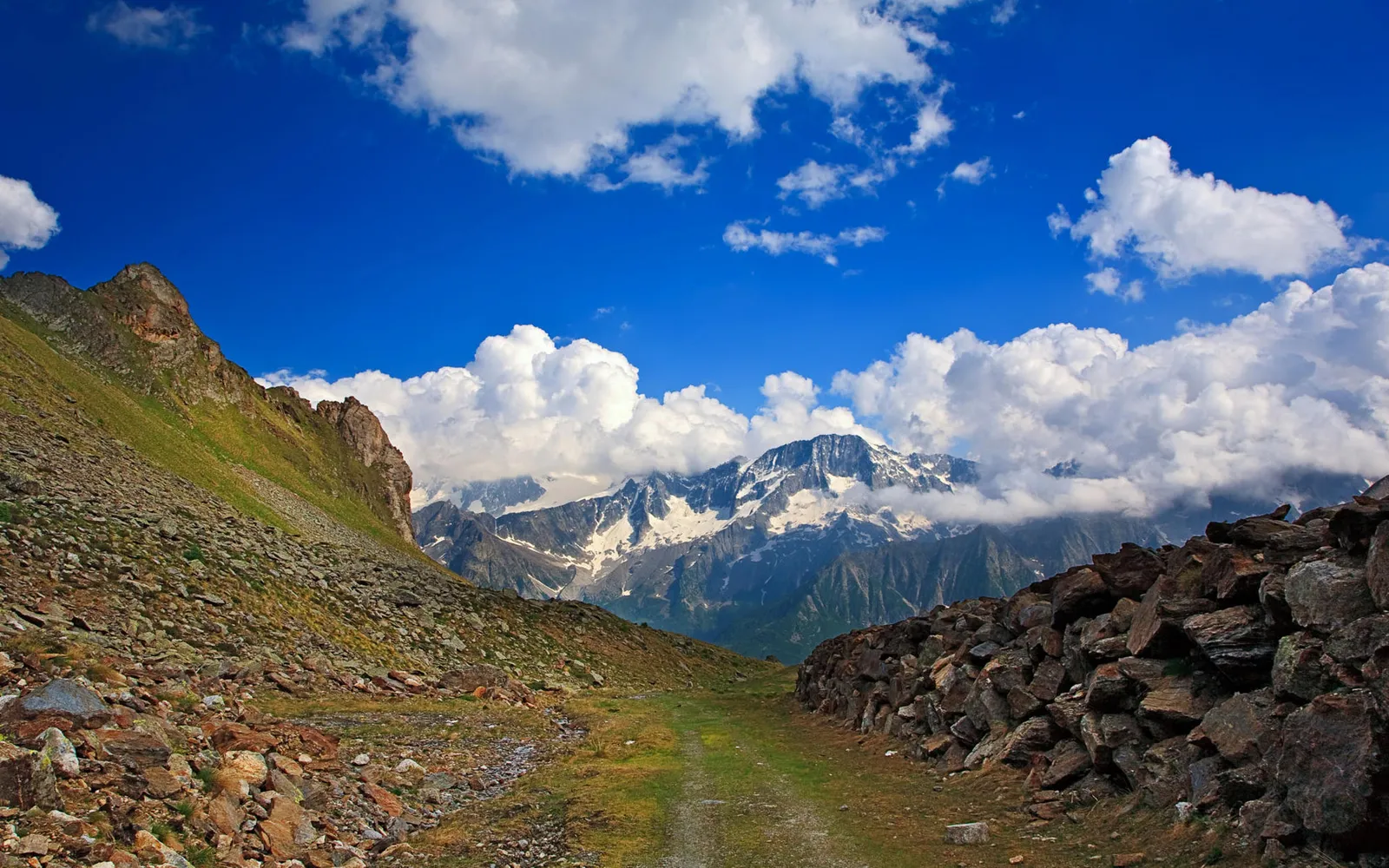 A rocky mountain path leads towards snow-capped peaks under a vibrant blue sky with fluffy white clouds.