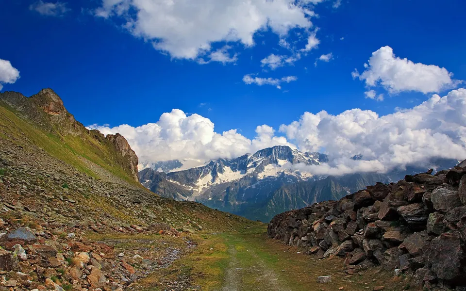 A rocky mountain path leads towards snow-capped peaks under a vibrant blue sky with fluffy white clouds.