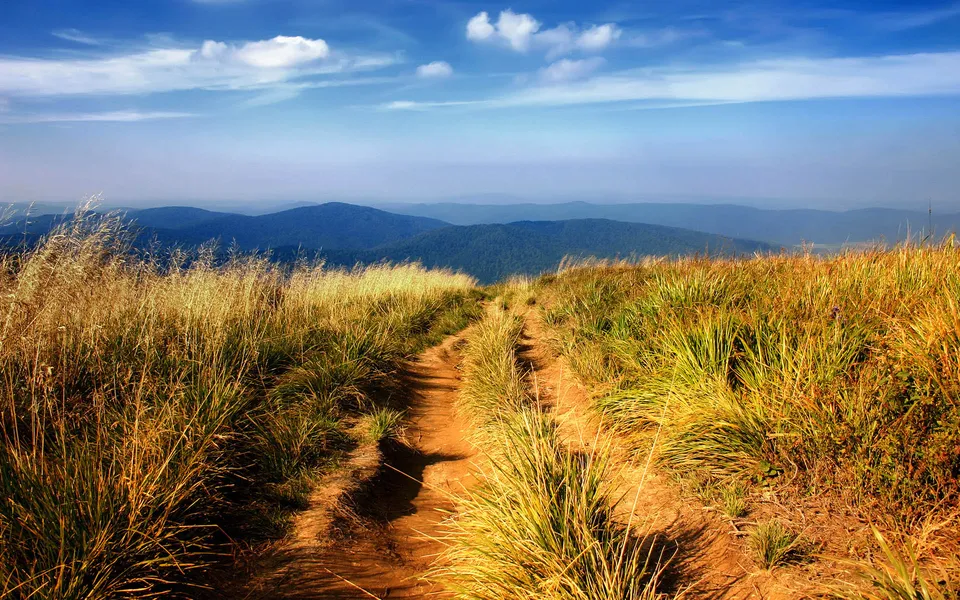 A dirt path winds through dry, golden grass up a mountain slope with blue hills in the distance.