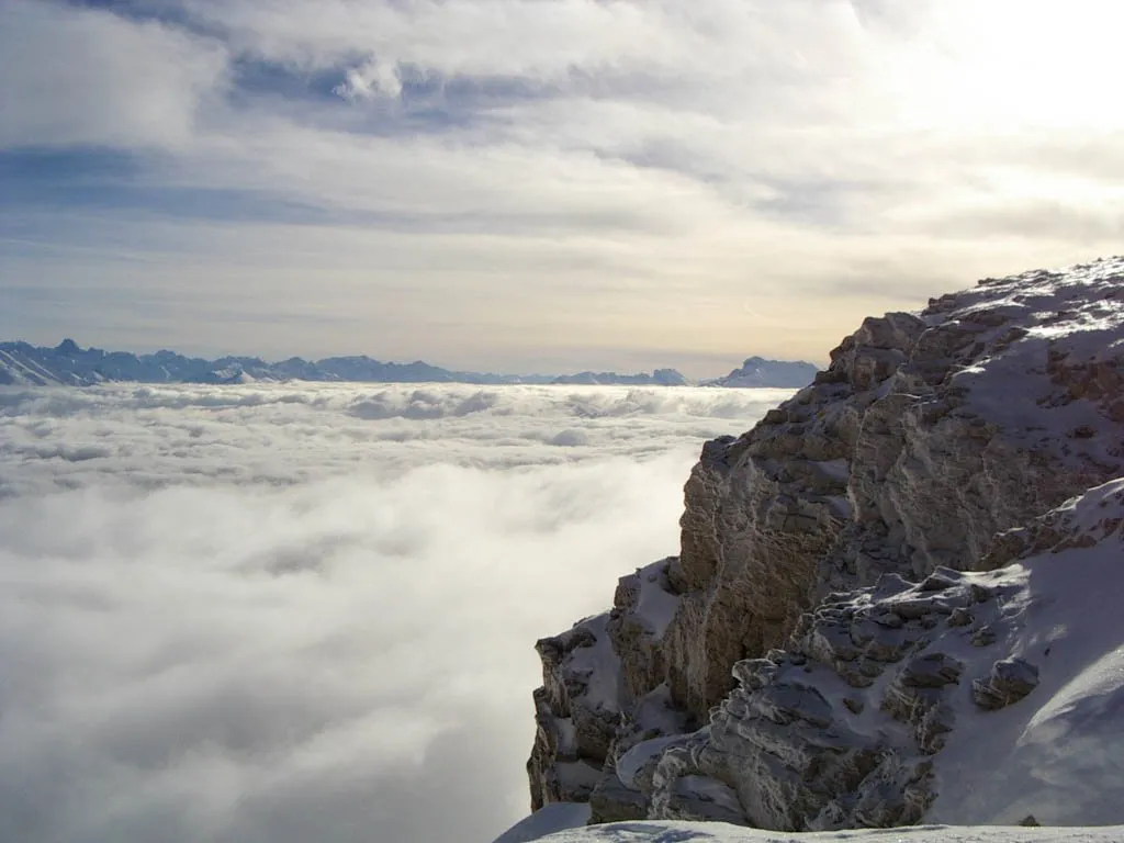 A rocky mountain cliff covered in snow overlooks a vast expanse of white clouds below.