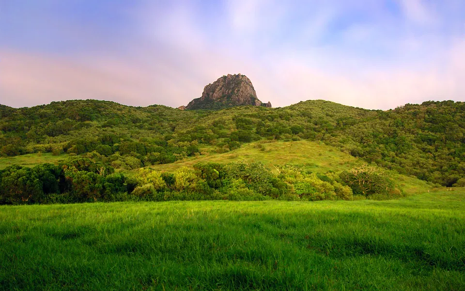 A rugged mountain peak rises above lush green hills and a foreground of vibrant grass under a soft twilight sky.