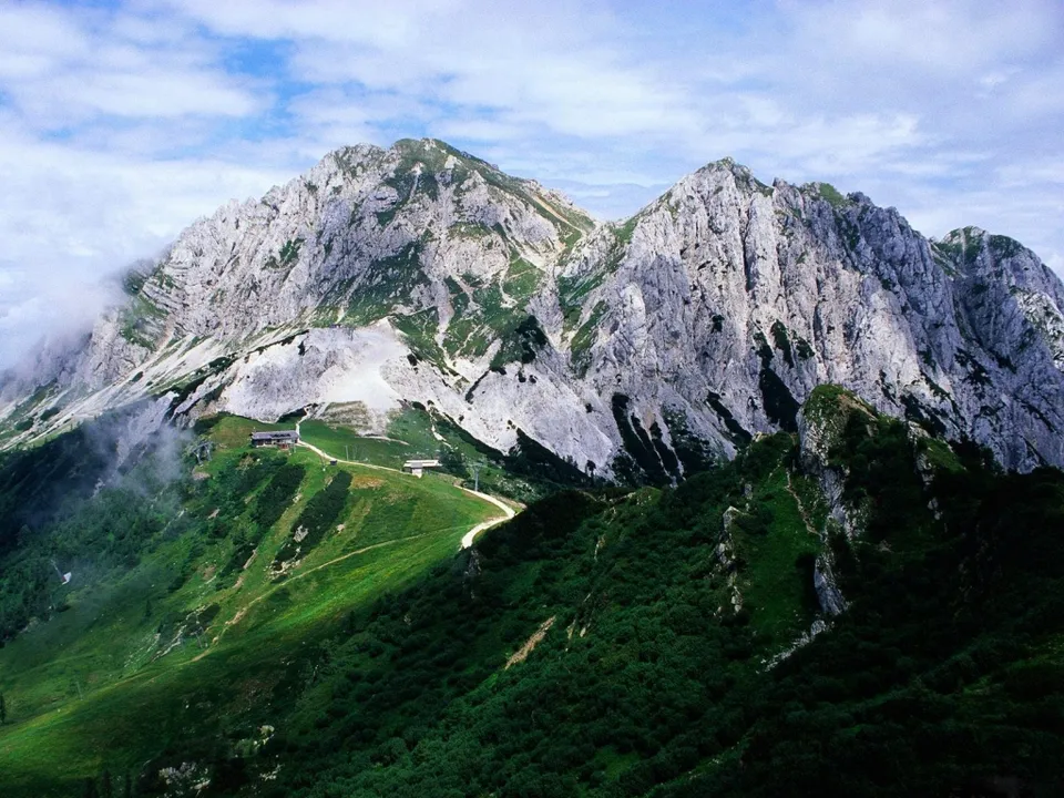 A wide view of rugged, rocky mountain peaks with green slopes and a winding path leading to buildings.
