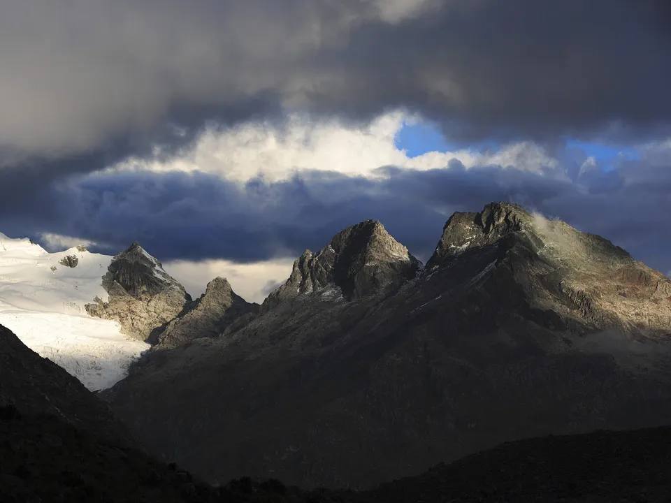 Jagged mountain peaks, one covered in snow, under a dramatic sky with dark clouds and patches of blue.