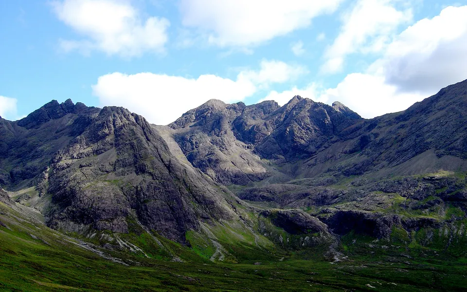 Jagged, dark grey mountain peaks rise steeply, with green grassy slopes in the foreground under a blue sky with white clouds.
