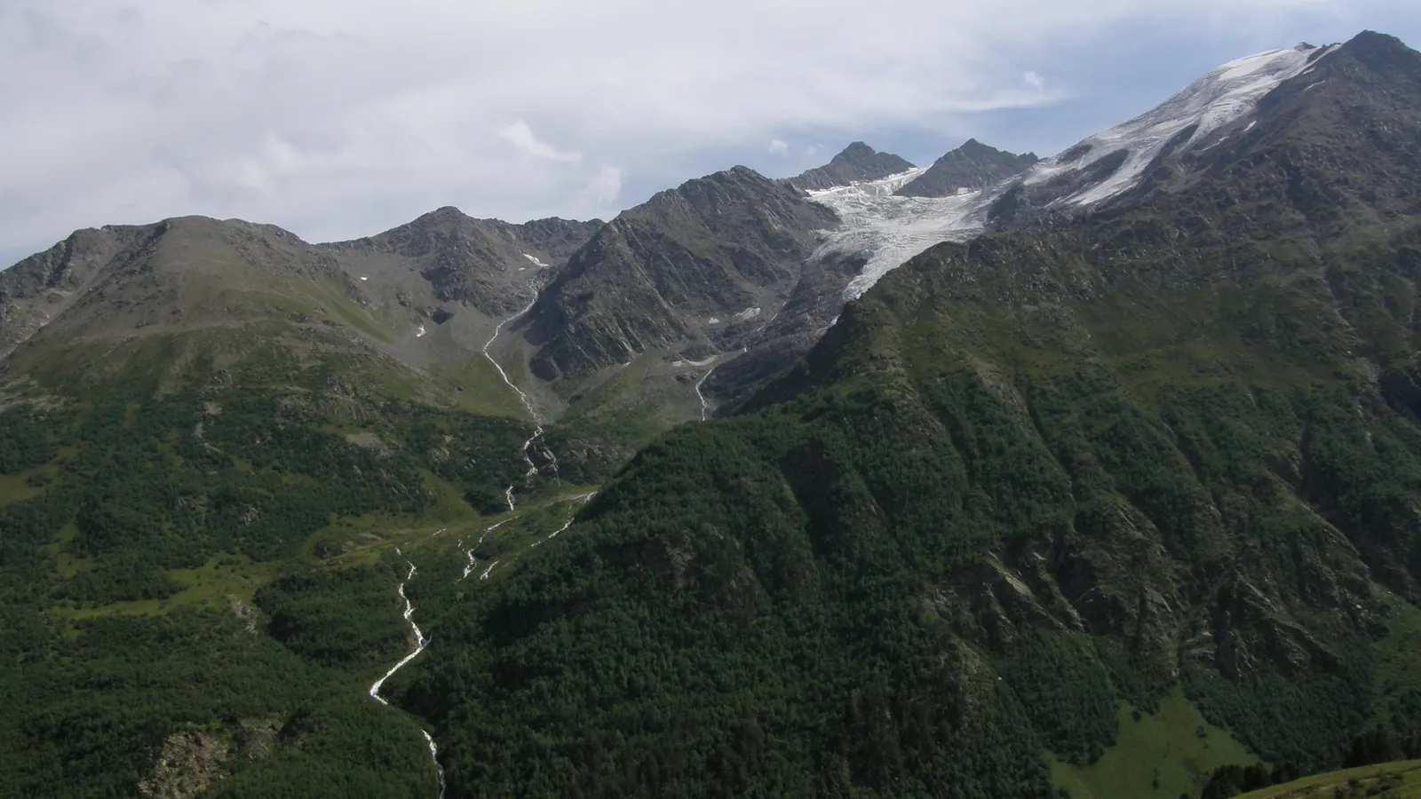 A wide view of rugged mountain peaks, some covered in snow and ice, with green forested slopes and a winding river below.