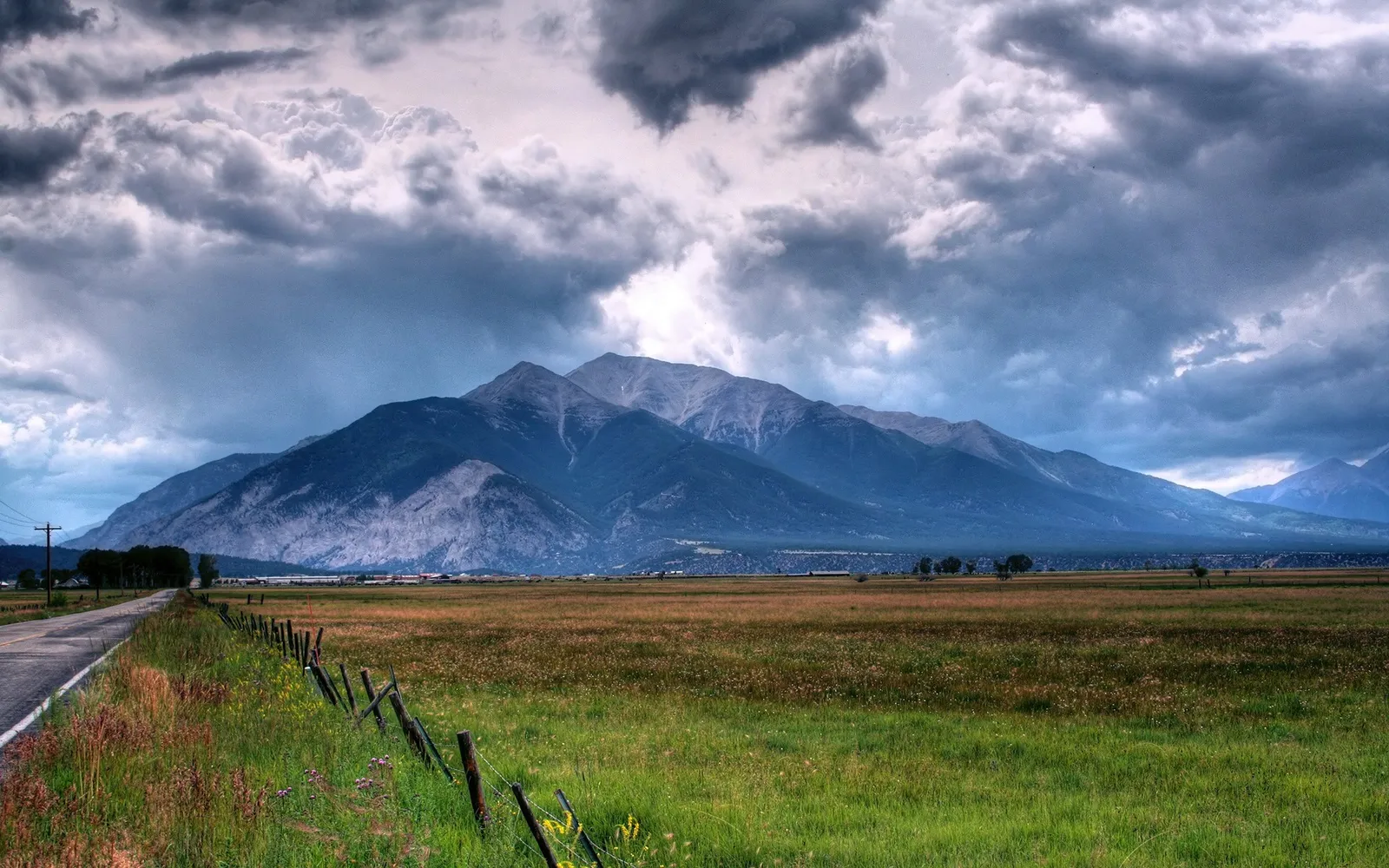 A vast mountain range under a dramatic, cloudy sky, with a field and road in the foreground.