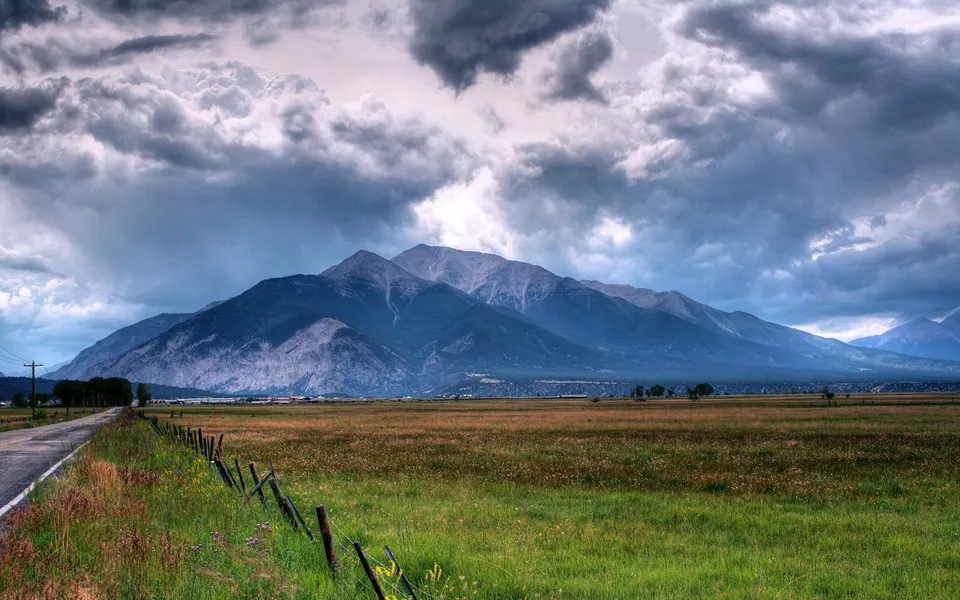 A vast mountain range under a dramatic, cloudy sky, with a field and road in the foreground.