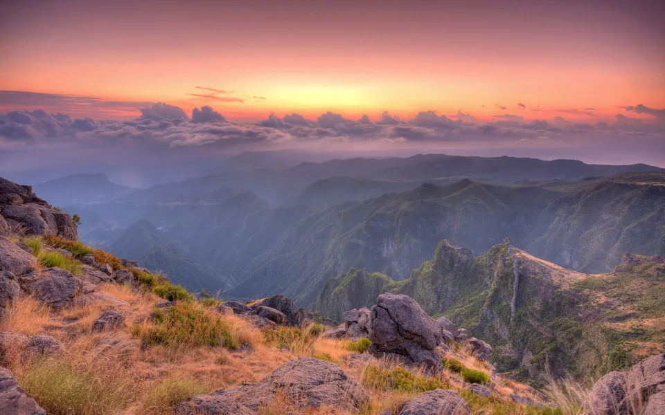 A panoramic view of layered mountain ranges fading into the distance under a vibrant sunset sky with clouds.