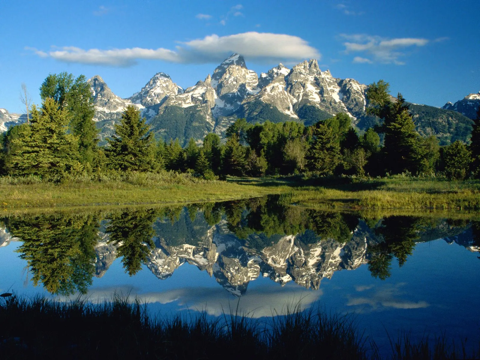 Snow-capped mountains reflected perfectly in a still lake, with green trees and blue sky.