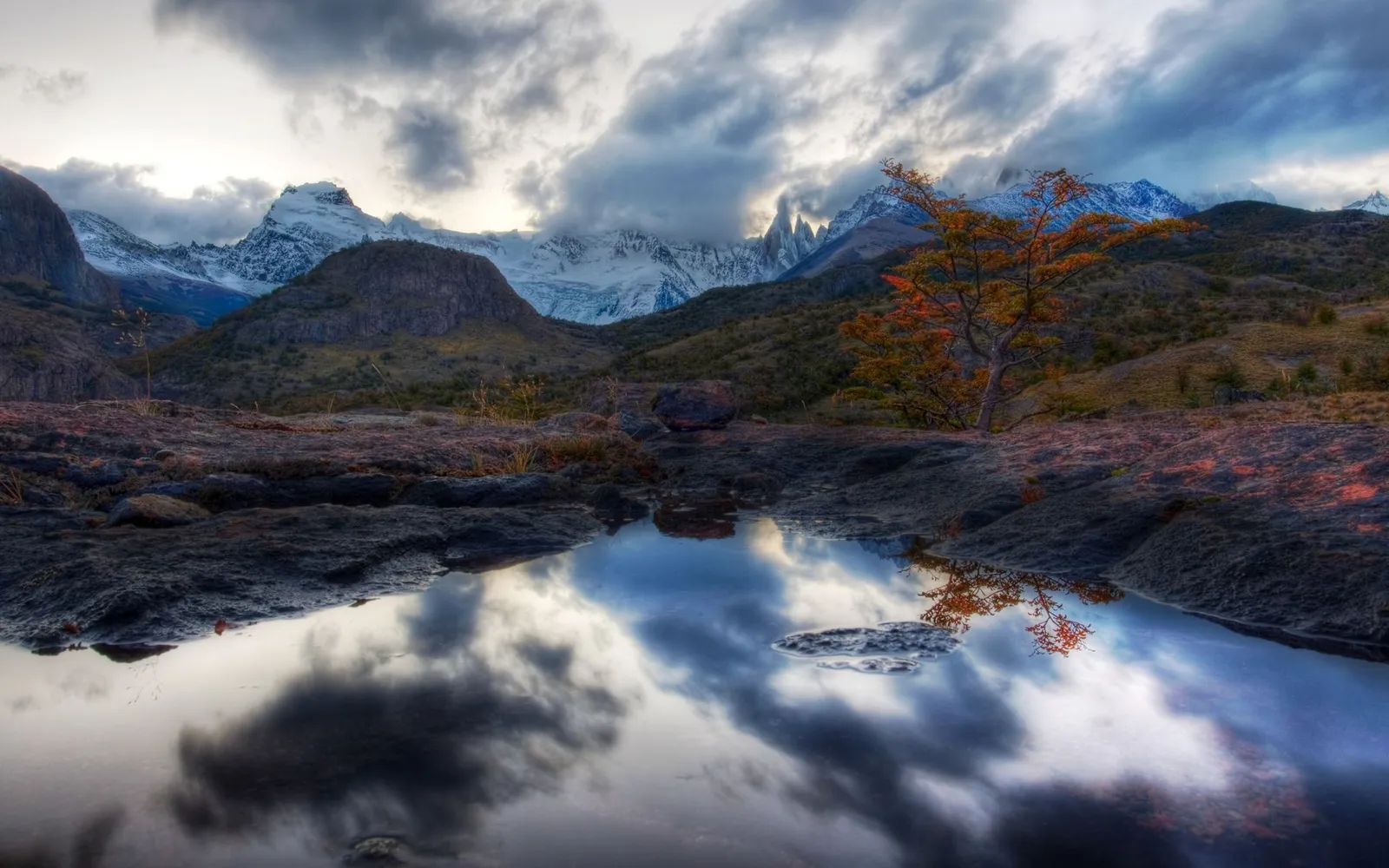 A dramatic mountain range with snow-capped peaks is reflected in a still puddle in the foreground.