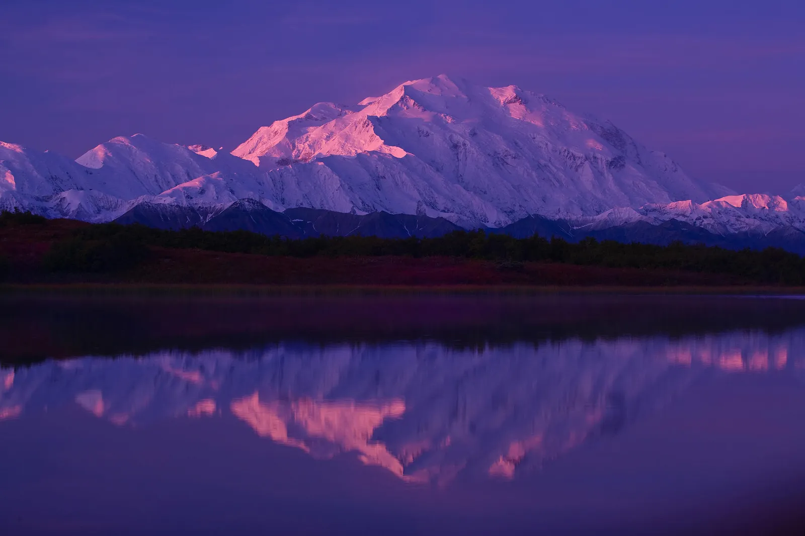 A snow-capped mountain range reflects in a calm lake during a purple and pink sunrise.
