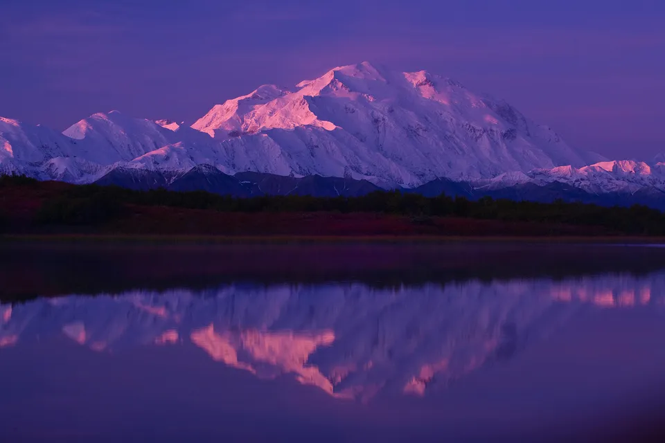 A snow-capped mountain range reflects in a calm lake during a purple and pink sunrise.