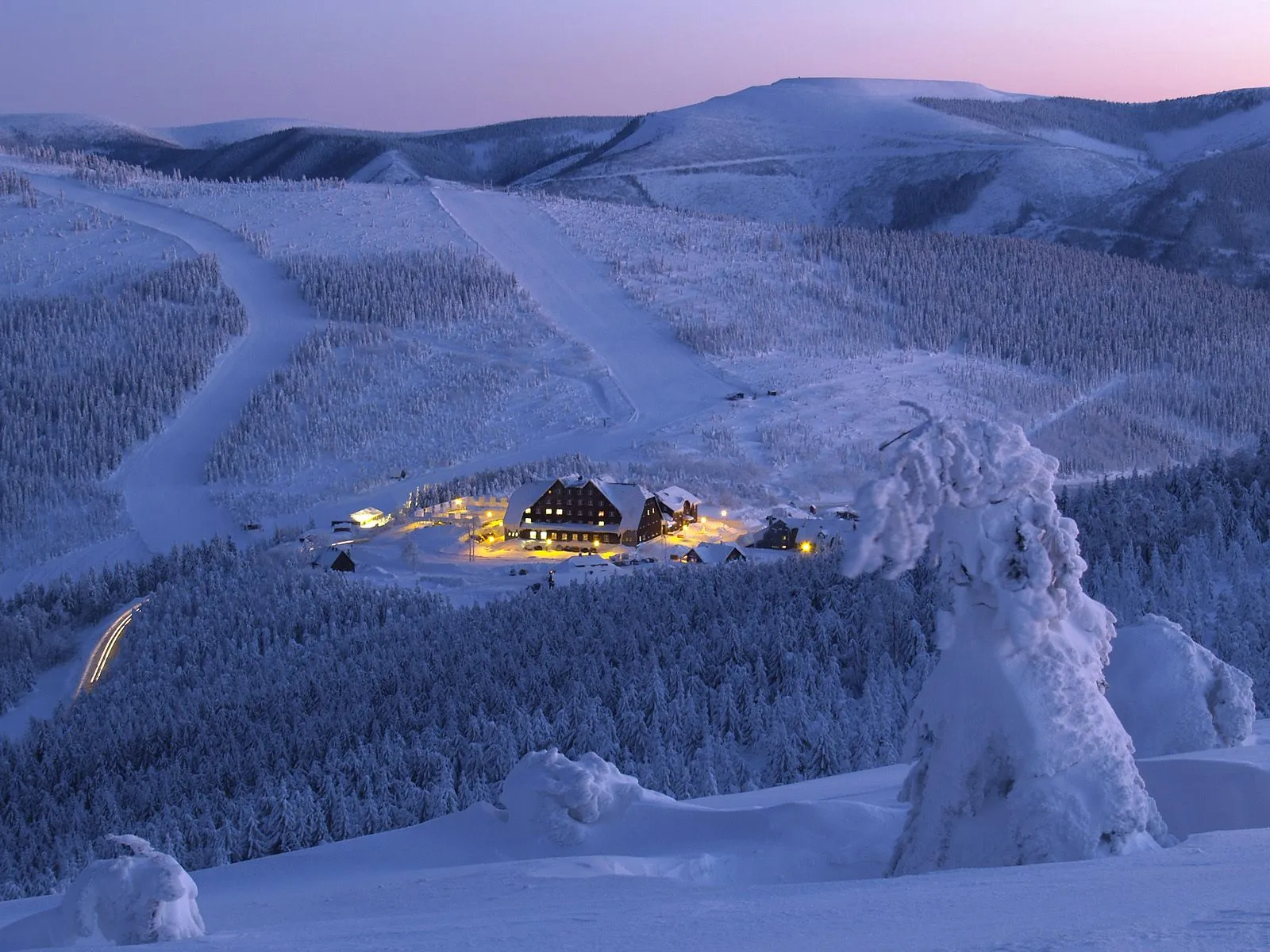 A mountain resort with lights on is nestled in a snow-covered forest, with ski slopes visible on the hills.