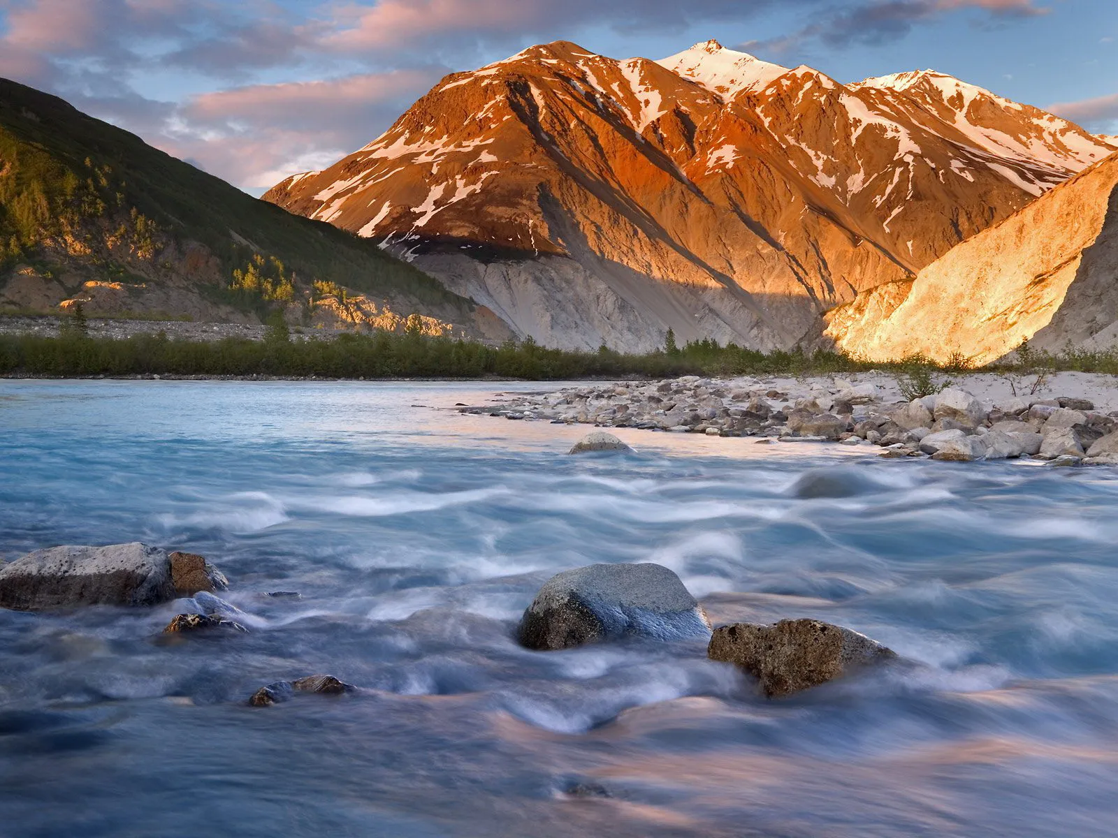 A wide, fast-flowing river in the foreground with snow-capped mountains bathed in golden light in the background.
