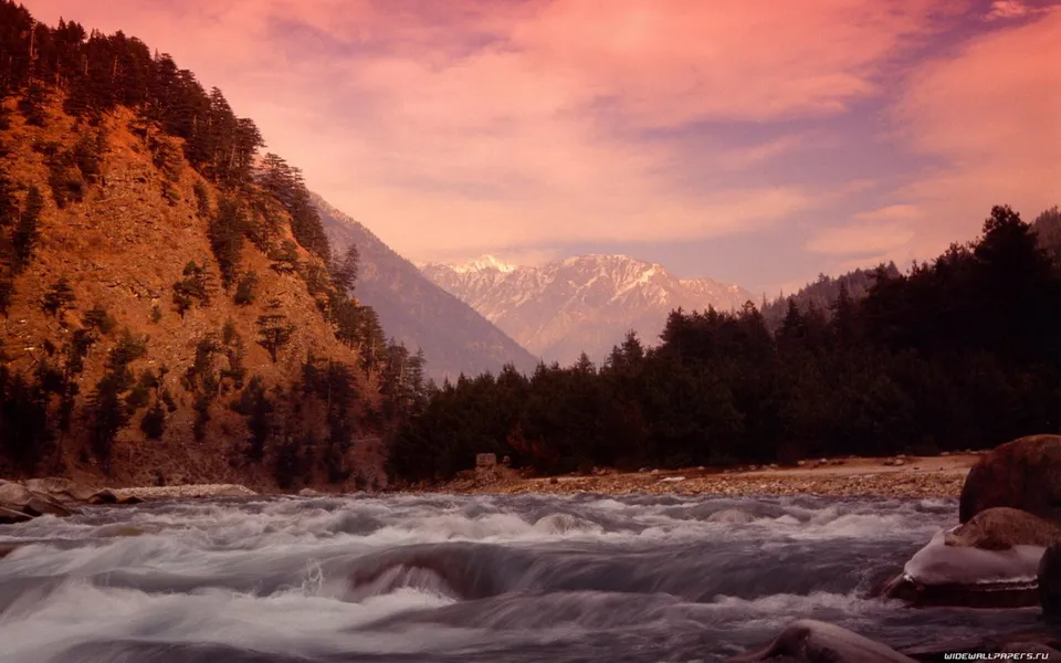 A wide, fast-flowing river in the foreground, with a steep, tree-covered mountain slope on the left and distant snow-capped mountains under a pink sky.