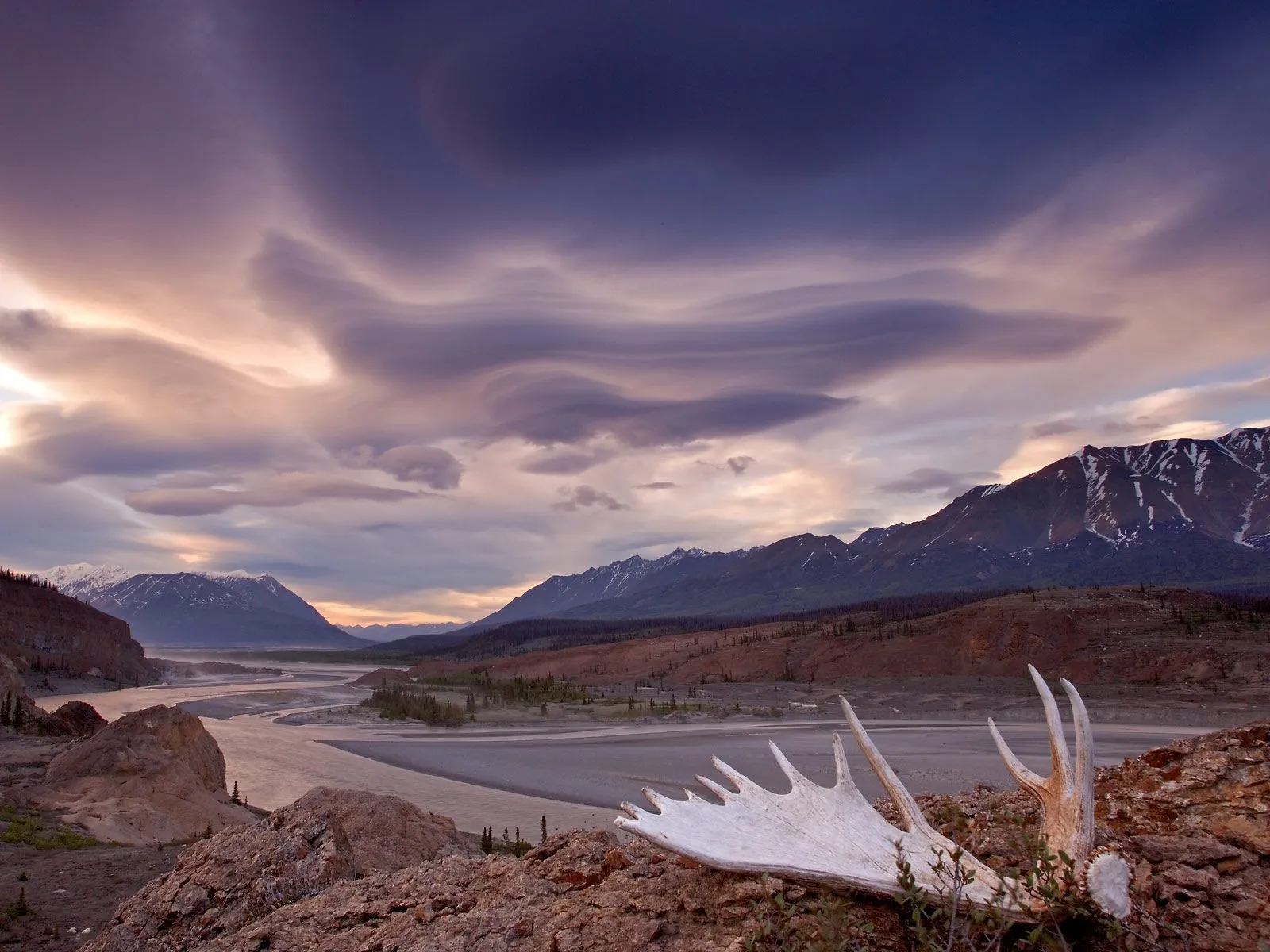 A wide river valley with mountains in the background under a dramatic, cloudy sky at sunset.
