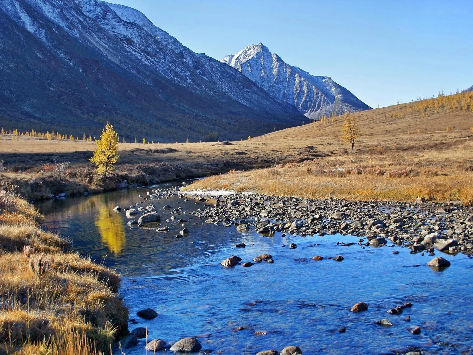 A clear blue stream flows through a rocky bed in a valley with mountains in the background.