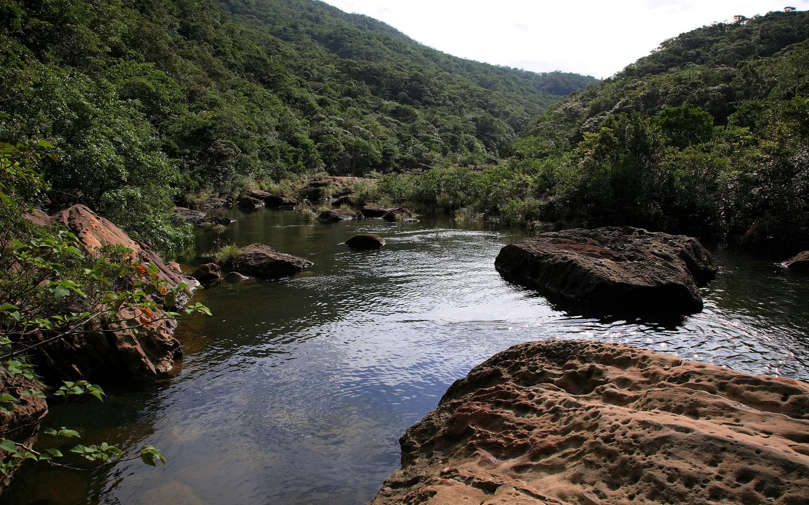 A wide, clear stream flows through a valley surrounded by dense green hills and large rocks.