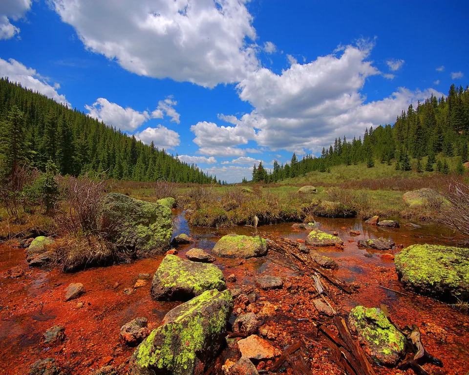 A shallow stream flows through a rocky area with red-colored stones and moss-covered boulders, leading towards a pine-covered mountainside under a blue sky with white clouds.