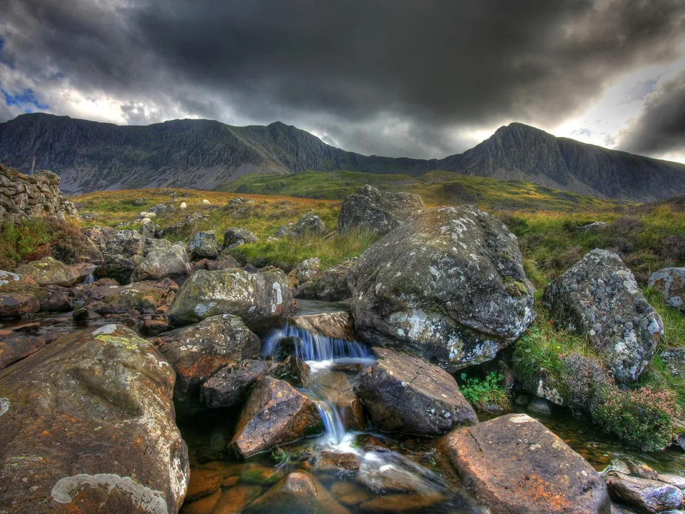 A rocky stream flows through a mountain landscape with dramatic clouds overhead.