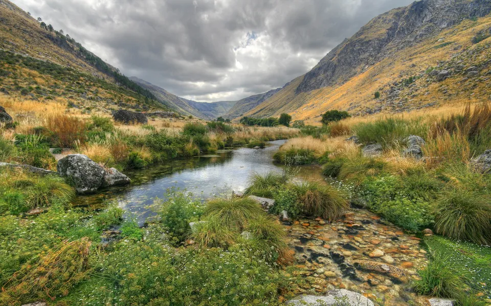 A clear mountain stream flows through a valley with grassy slopes and cloudy skies.