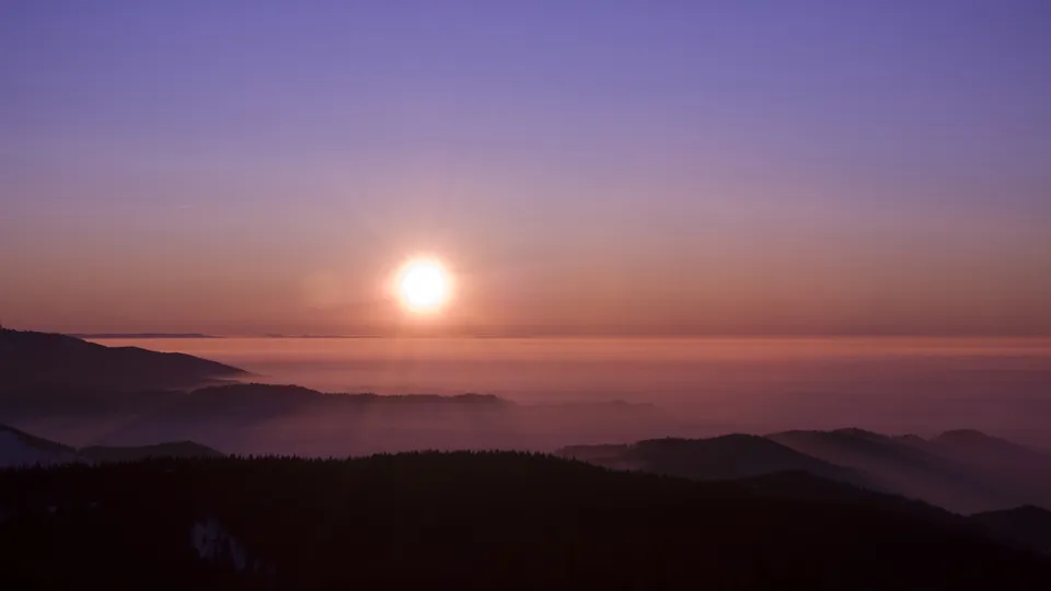 The sun rises over layers of misty mountains, casting a warm glow.