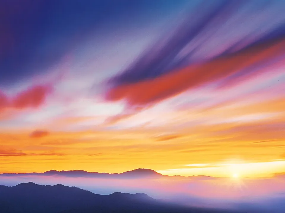 A vibrant sunrise illuminates the sky with orange, pink, and purple hues over silhouetted mountain ranges shrouded in mist.