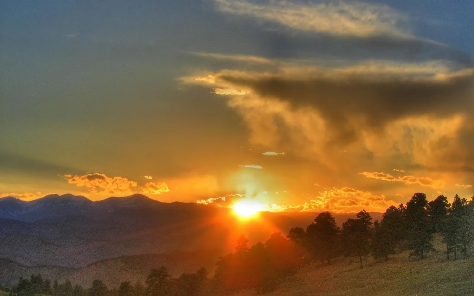 The sun sets behind a mountain range, casting golden rays through clouds and illuminating pine trees.