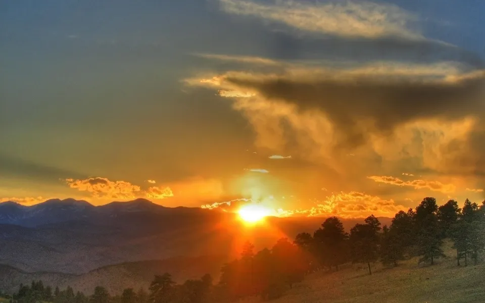 The sun sets behind a mountain range, casting golden rays through clouds and illuminating pine trees.