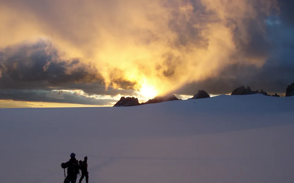 Two hikers silhouetted against a vast, snow-covered landscape with dramatic clouds at sunset.