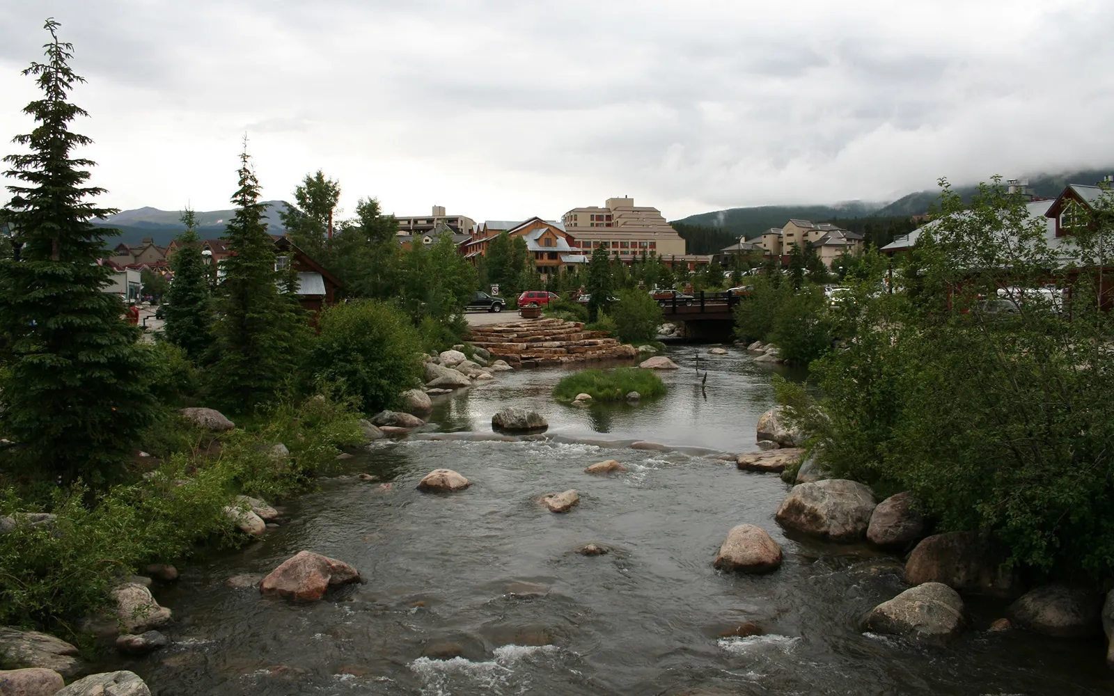 A wide river with rocks flows through a mountain town with buildings and trees under a cloudy sky.