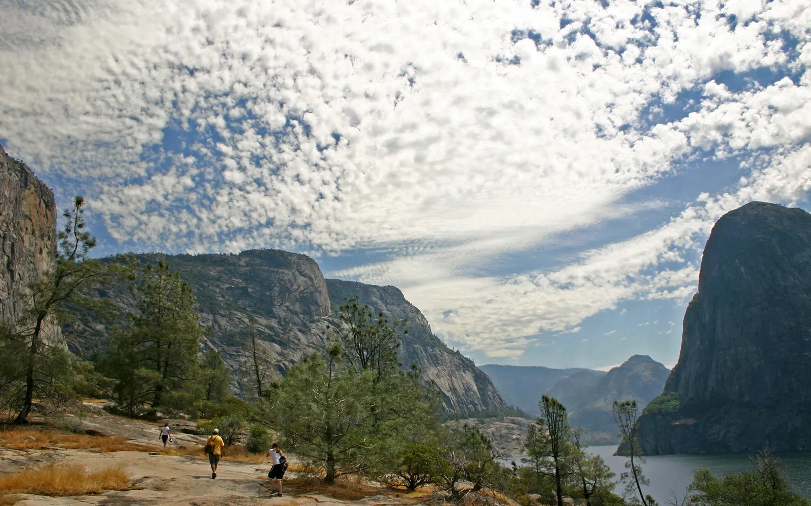 Three hikers walk along a rocky path with large granite cliffs and a cloudy blue sky.