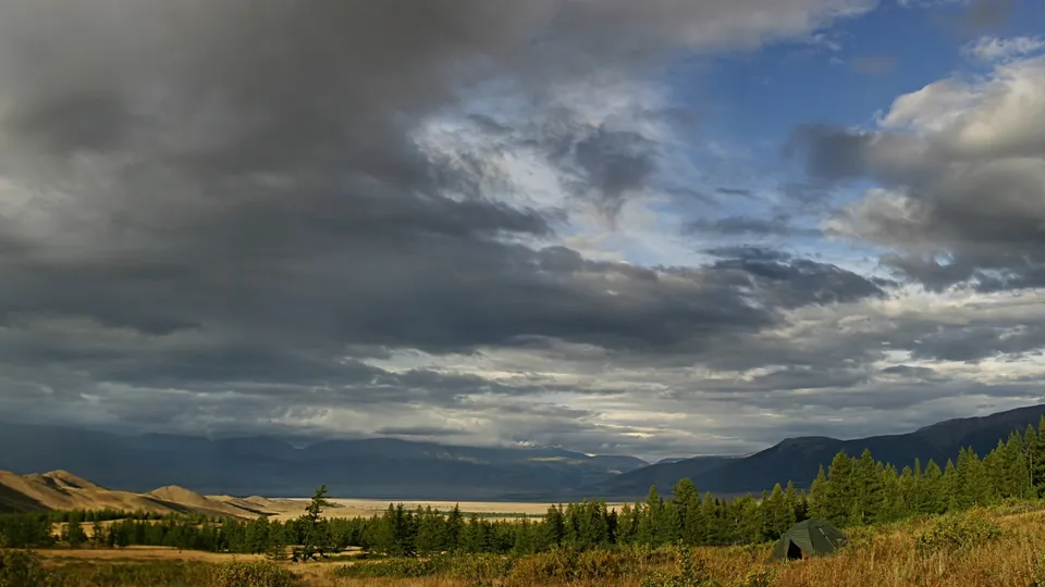 A wide landscape view of a mountain valley with a tent in the foreground and dramatic clouds in the sky.