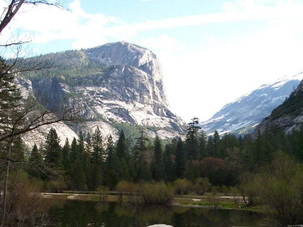 A wide mountain valley with a calm lake in the foreground and a large granite cliff face on the left.