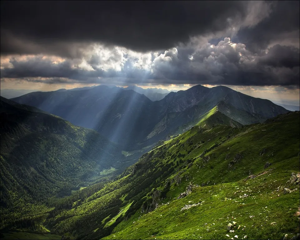 Sunbeams stream through dark clouds onto a lush green mountain valley and peaks.