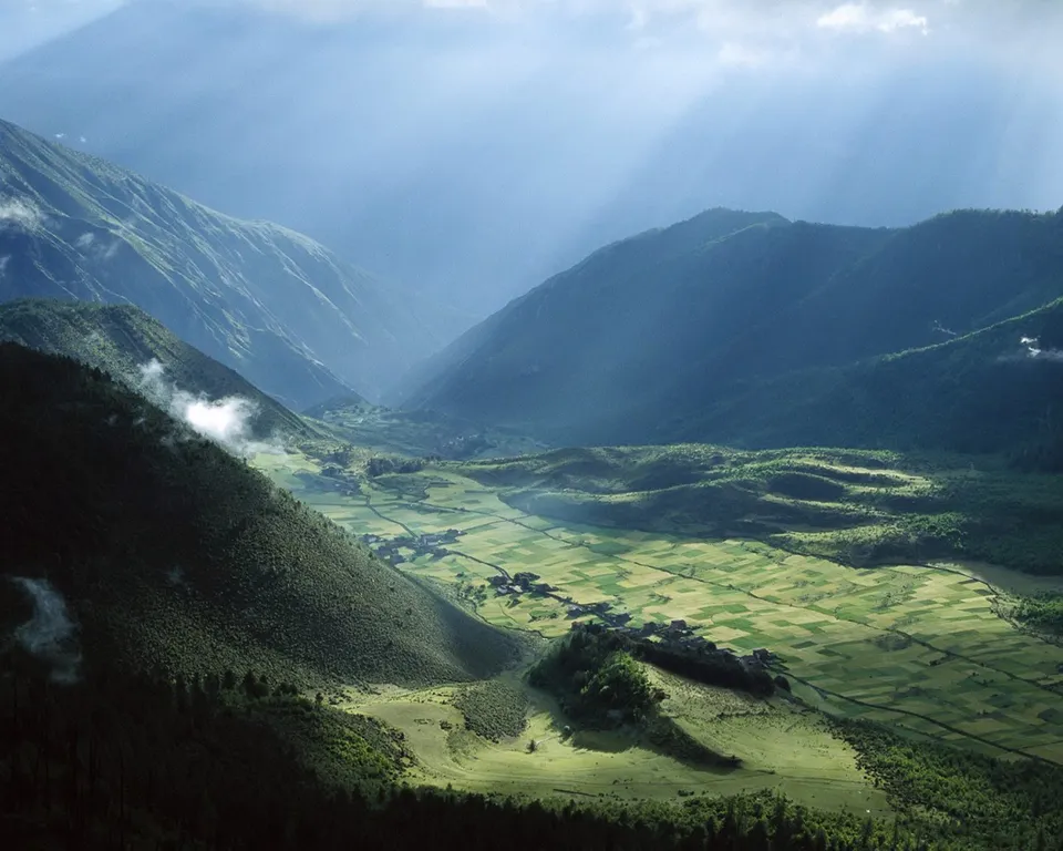 A wide valley with patchwork fields is illuminated by dramatic sunbeams breaking through clouds above mountains.