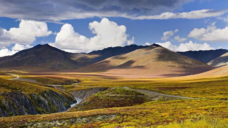 A winding road leads through a valley of colorful tundra towards distant mountains under a sky with dramatic clouds.