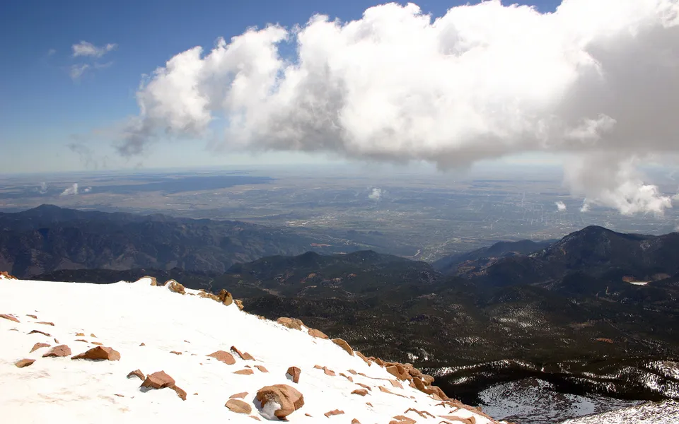 A vast landscape viewed from a snowy mountain peak, with clouds overhead and a distant valley below.