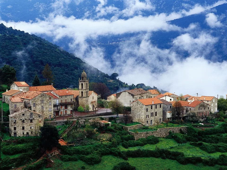 A stone village with a church tower sits on a green hillside, with misty clouds obscuring the mountain peaks above.