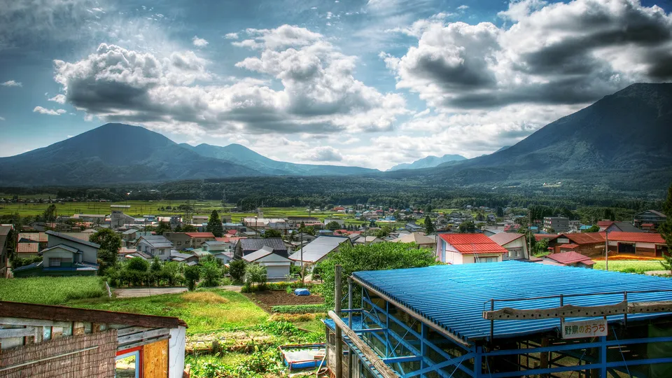 A wide shot of a village with mountains in the background and a cloudy sky.