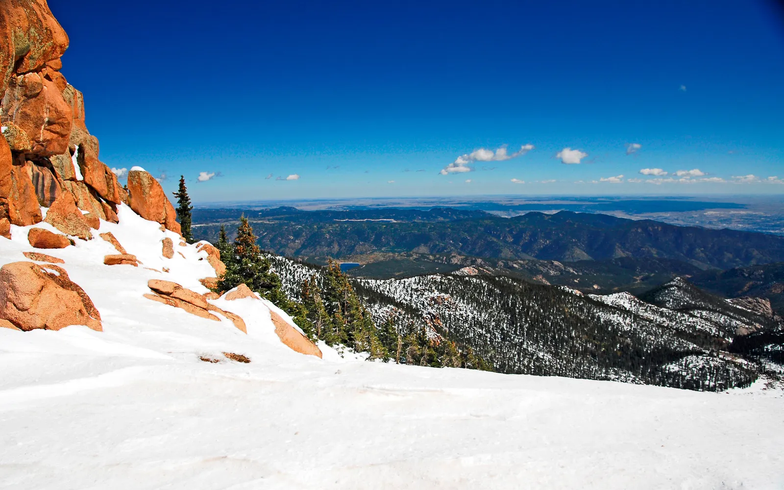 A wide view of snow-covered mountains and pine trees under a bright blue sky with scattered clouds.
