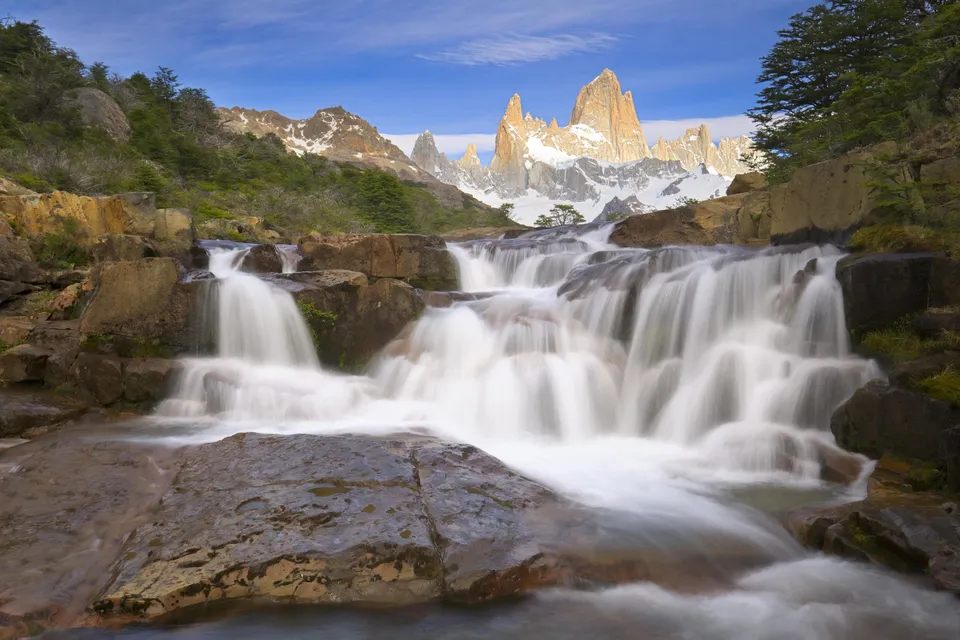 A multi-tiered waterfall cascades over rocks in the foreground, with snow-capped, jagged mountain peaks in the background under a blue sky.