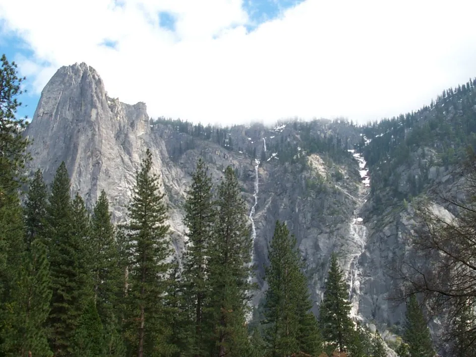 A tall, thin waterfall flows down a steep, rocky mountain face, surrounded by pine trees.