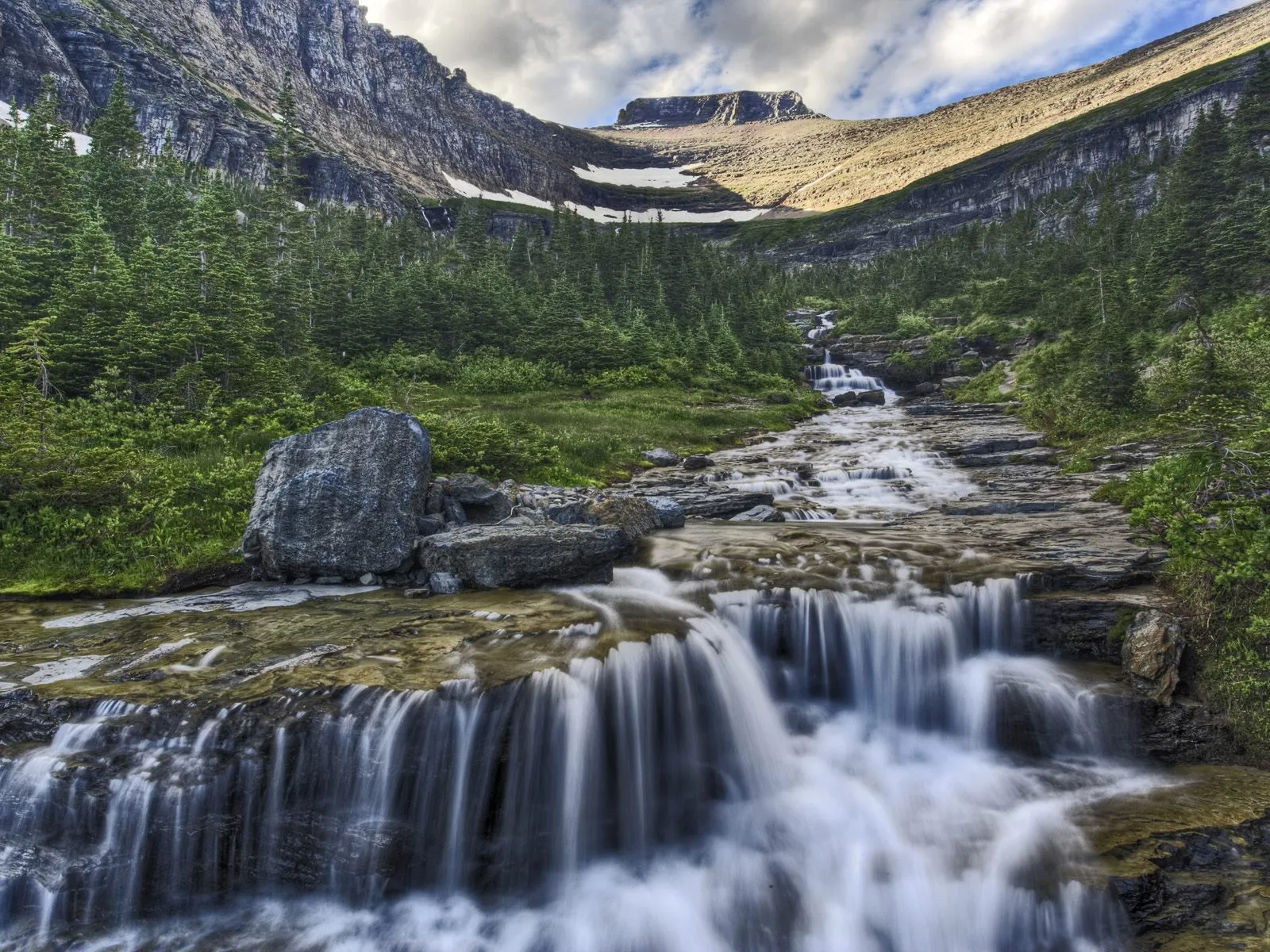 A multi-tiered waterfall flows down a rocky mountain slope surrounded by pine trees.