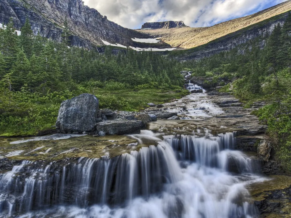 A multi-tiered waterfall flows down a rocky mountain slope surrounded by pine trees.