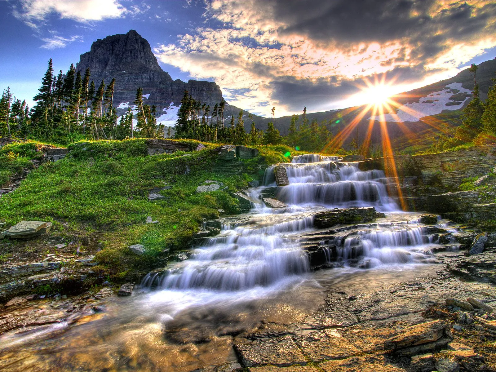 A multi-tiered waterfall cascades down rocky terrain with a mountain peak and sunburst in the background.