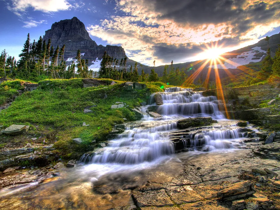 A multi-tiered waterfall cascades down rocky terrain with a mountain peak and sunburst in the background.